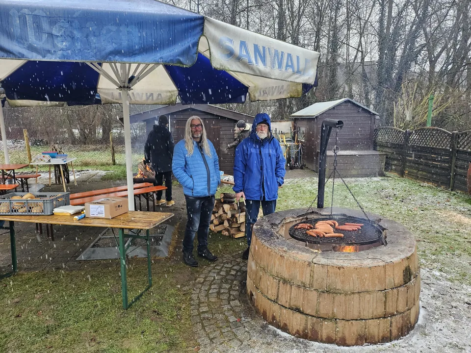 Würstchen auf dem Schwenkgrill über dem Feuer, zwei Helfer im Schneetreiben